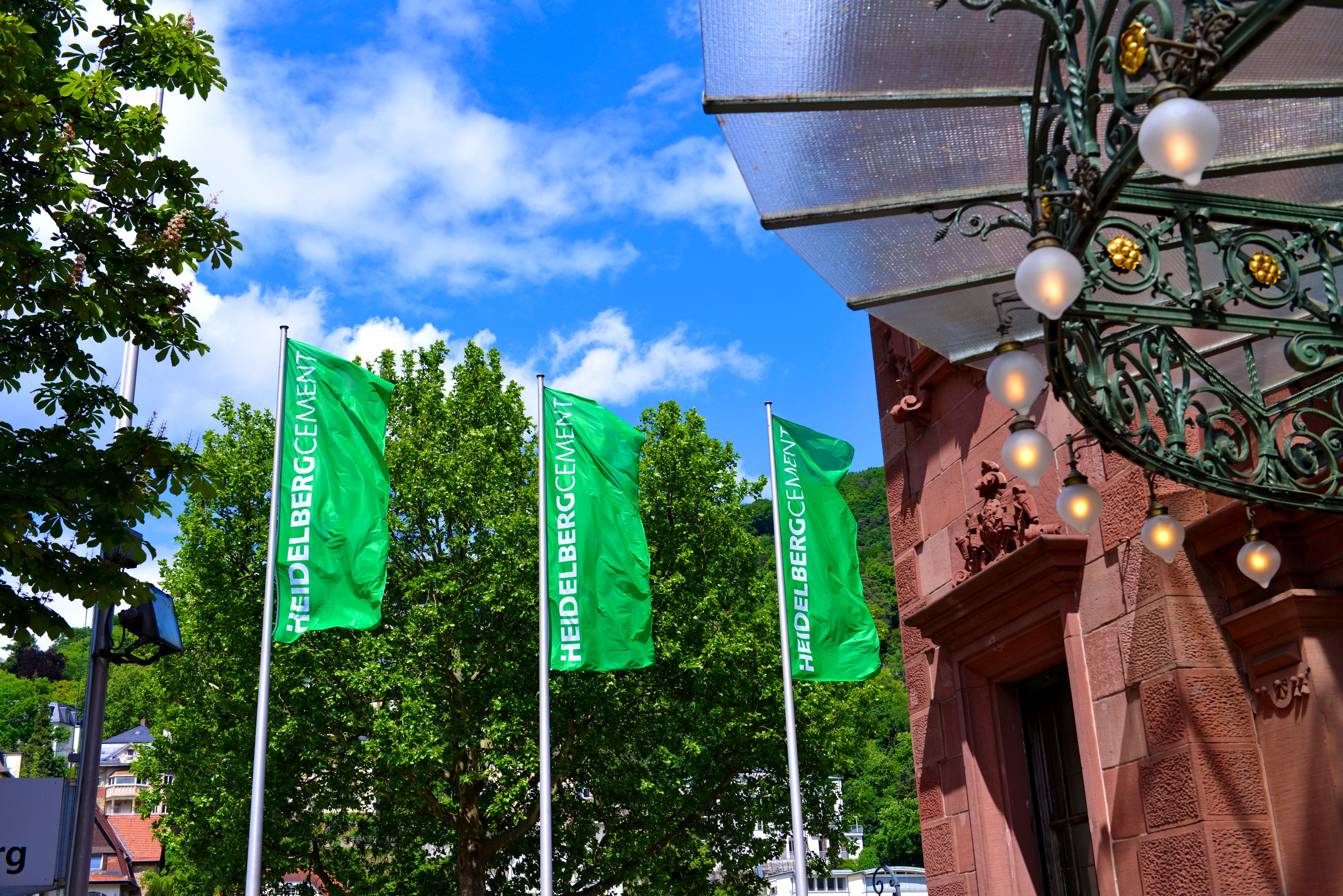 Three green flags with HeidelbergCement lettering next to a sandstone facade, in front of green trees