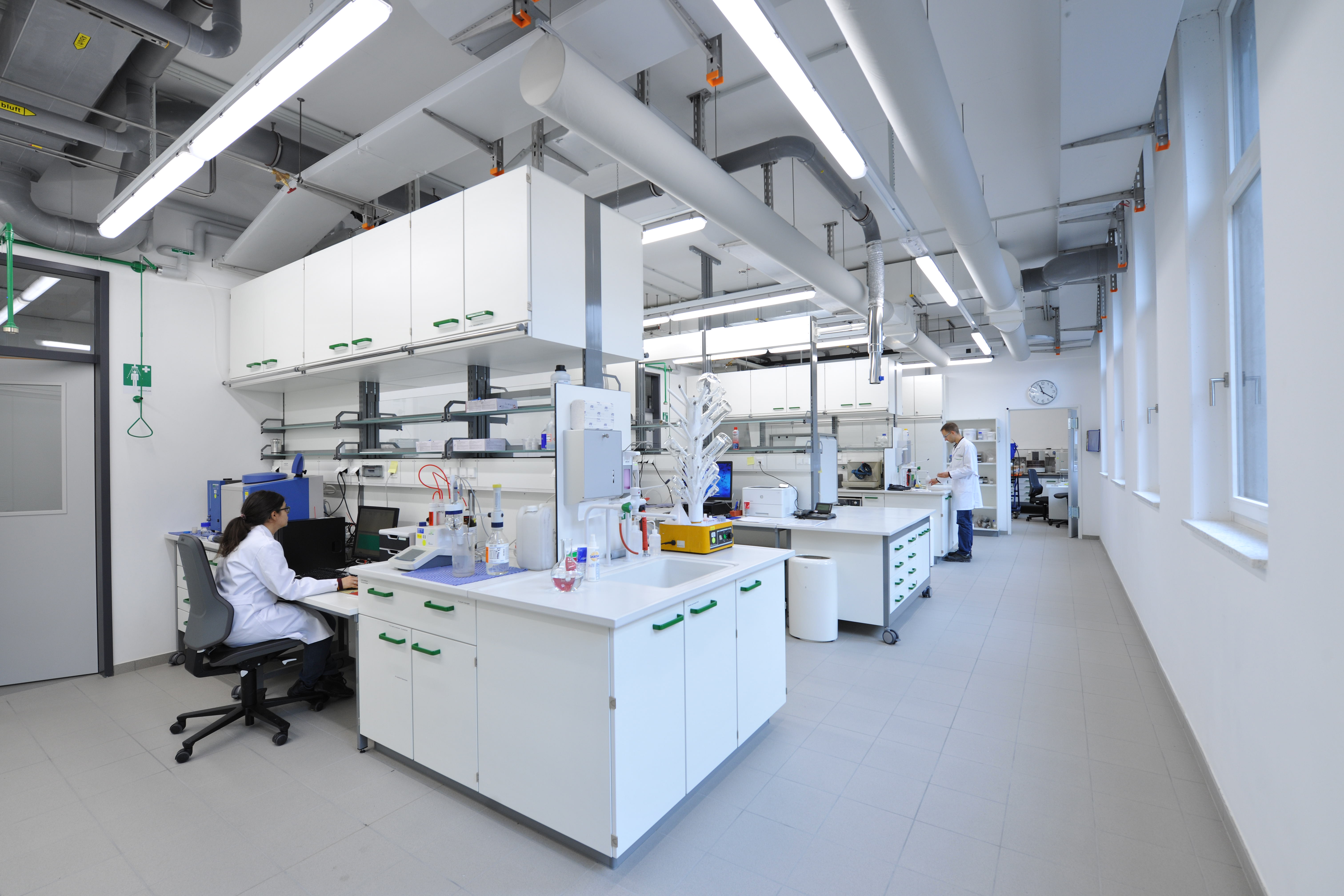 Labor mit Schreibtischen und Schränken, stehende und sitzende Personen in Laborkleidern