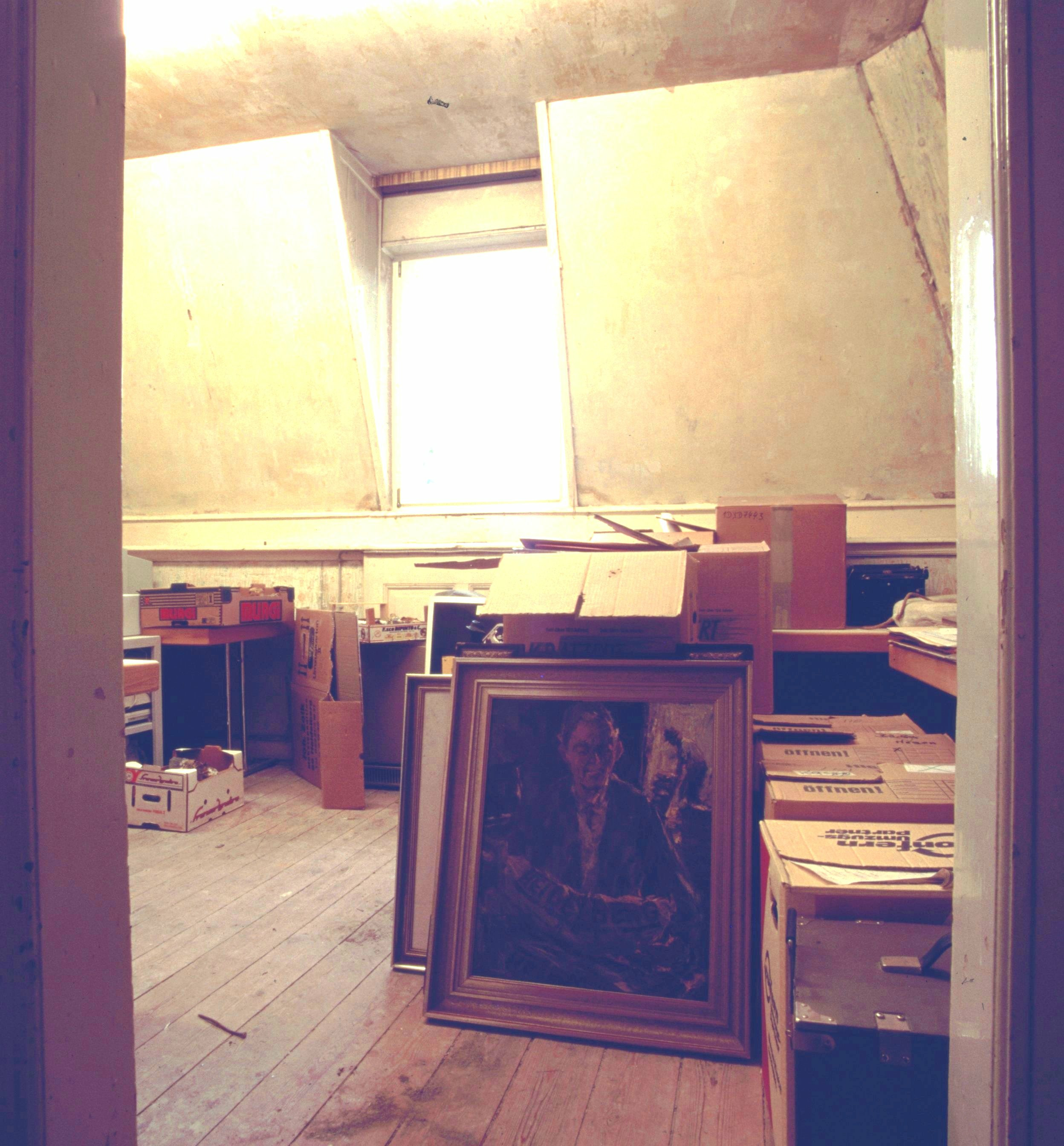 Karger Raum mit Holzboden, in dem Kisten und eingerahmte Bilder stehen