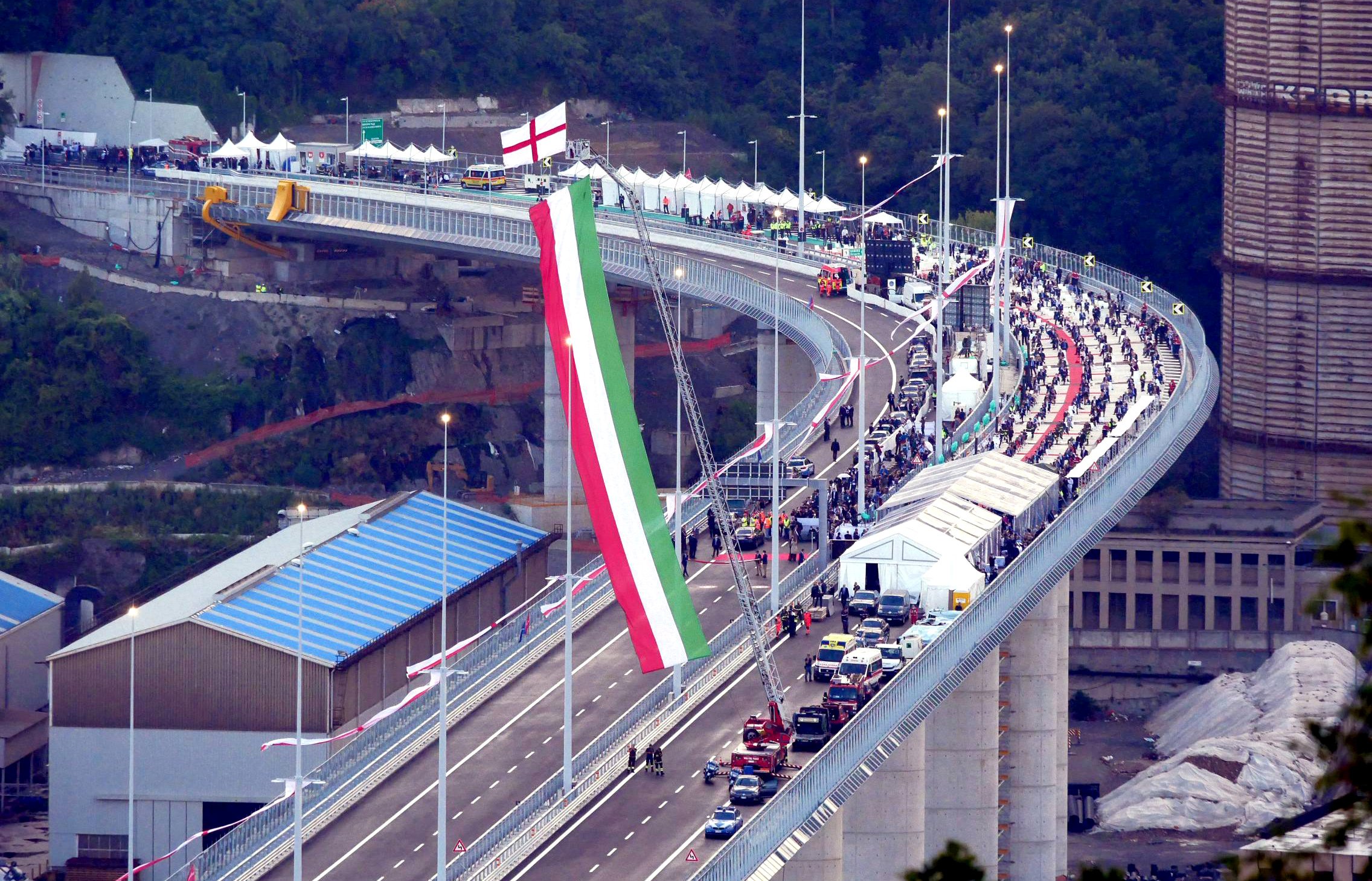 Eine geschwungene Brücke mit einer großen Menschenmenge darauf, die italienische Flagge wird von einem Kran gehisst, der an einem Feuerwehrauto befestigt ist