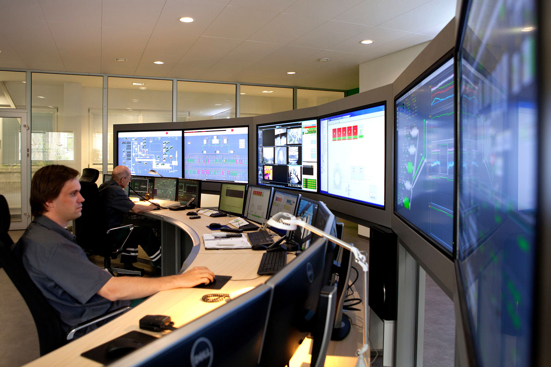 Three people sit in front of many control monitors